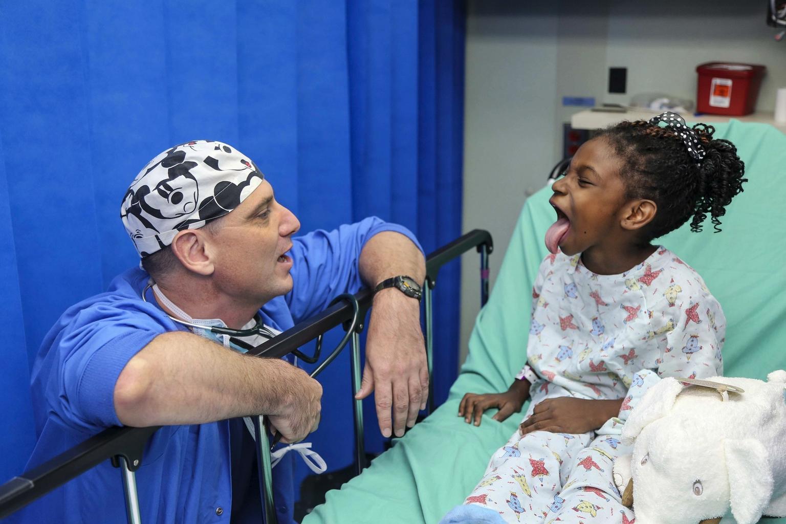 A clinician in a Mickey Mouse scrub cap sharing a laugh with a young patient at the bedside